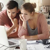 Serious man and woman sitting at kitchen table in front of open laptop computer, looking at screen with concentrated expression, focused on paying utility bills online. Family budget and finances